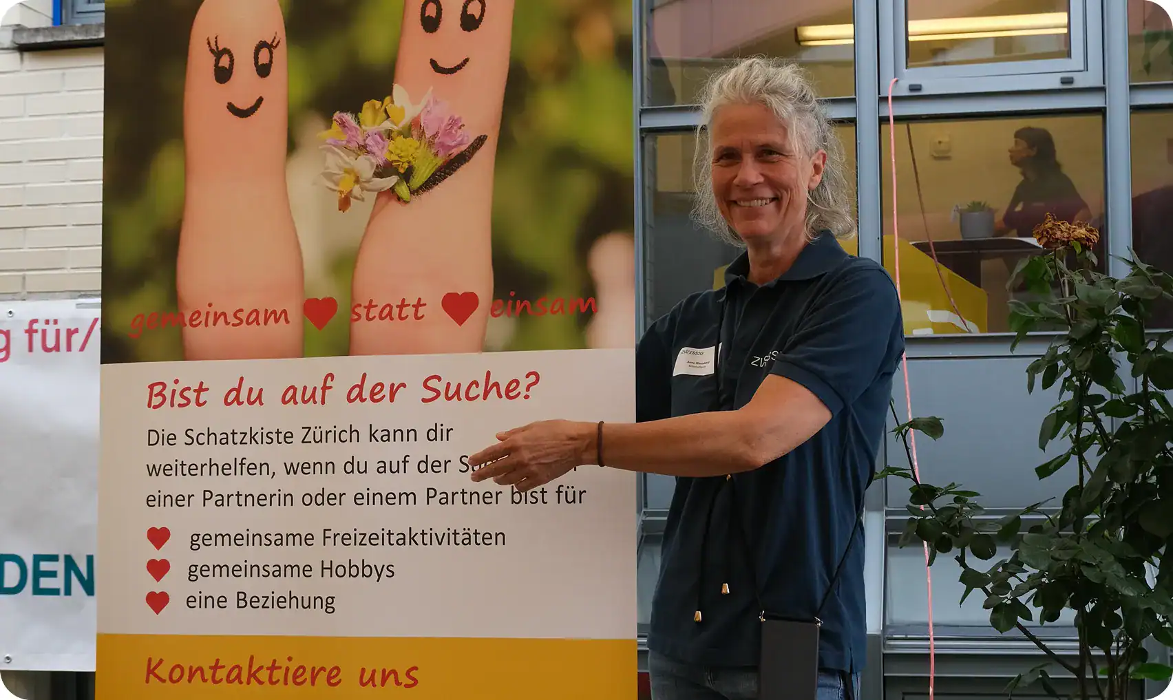 Eine Frau steht in einem Haus, lächelt und zeigt auf ein Banner mit zwei verzierten Fingern und deutschem Text, das über Beziehungen und soziale Aktivitäten informiert.