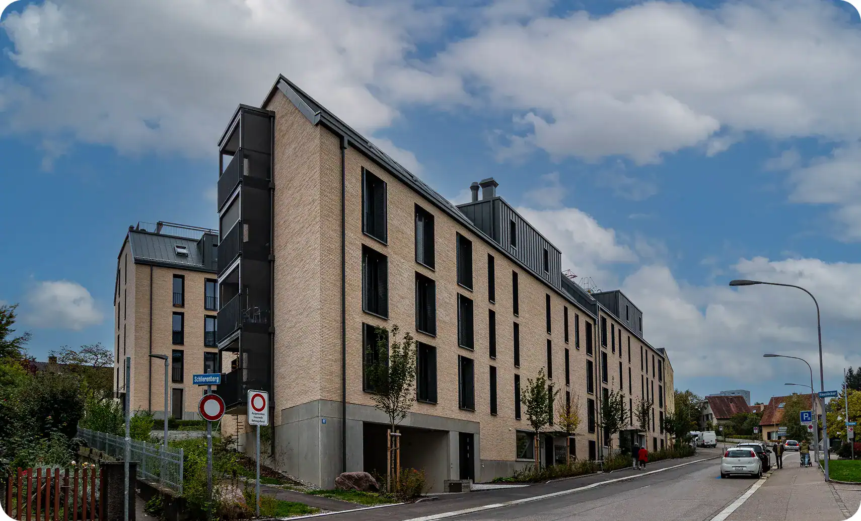 Modernes, mehrstöckiges Mehrfamilienhaus mit beiger Backsteinfassade und schwarzen Fensterrahmen an einer abschüssigen städtischen Straße unter einem teilweise bewölkten Himmel. Autos und Fußgänger sind zu sehen.