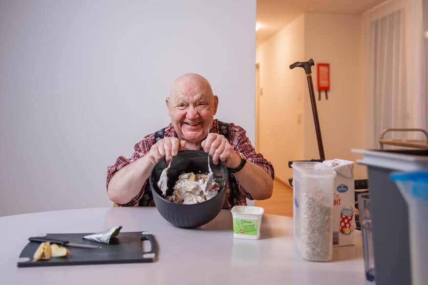 Ein älterer Mann sitzt an einem Tisch und rührt lächelnd Essen in einer Schüssel an. Auf dem Tisch stehen verschiedene Küchenutensilien und Zutaten, und in der Nähe ruht ein Stock.