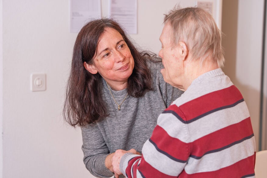 Eine Frau in einem grauen Pullover hält die Hand und lächelt einen älteren Mann an, der ein rot-grau gestreiftes Hemd in einem Innenraum trägt.