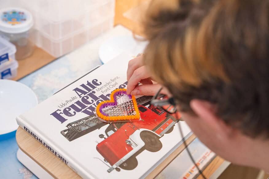 Eine Person legt ein perlenbesticktes, herzförmiges Kunstwerk auf ein Buch mit einem roten Feuerwehrauto auf dem Einband.