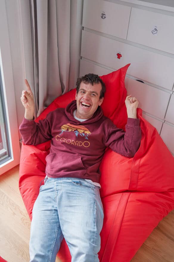 Mann in kastanienbrauner Colorado-Kapuzenjacke und Jeans, der auf einem roten Sitzsack sitzt, lächelt und mit beiden Händen nach oben zeigt.