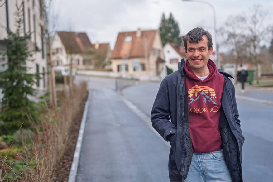 Ein Mann mit Kapuzenpulli und Jacke aus Colorado steht lächelnd in einer ruhigen Wohnstraße mit Häusern im Hintergrund.