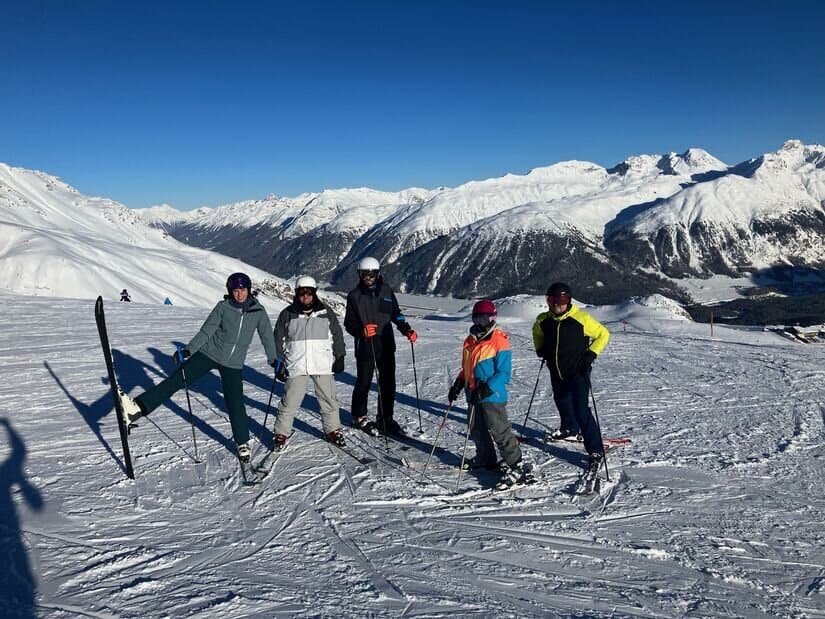 Fünf Personen in Skikleidung stehen mit Skistöcken auf einem verschneiten Berghang, umgeben von schneebedeckten Gipfeln unter einem strahlend blauen Himmel, und machen auf spenden für soziale Projekte in alpinen Gemeinden aufmerksam.