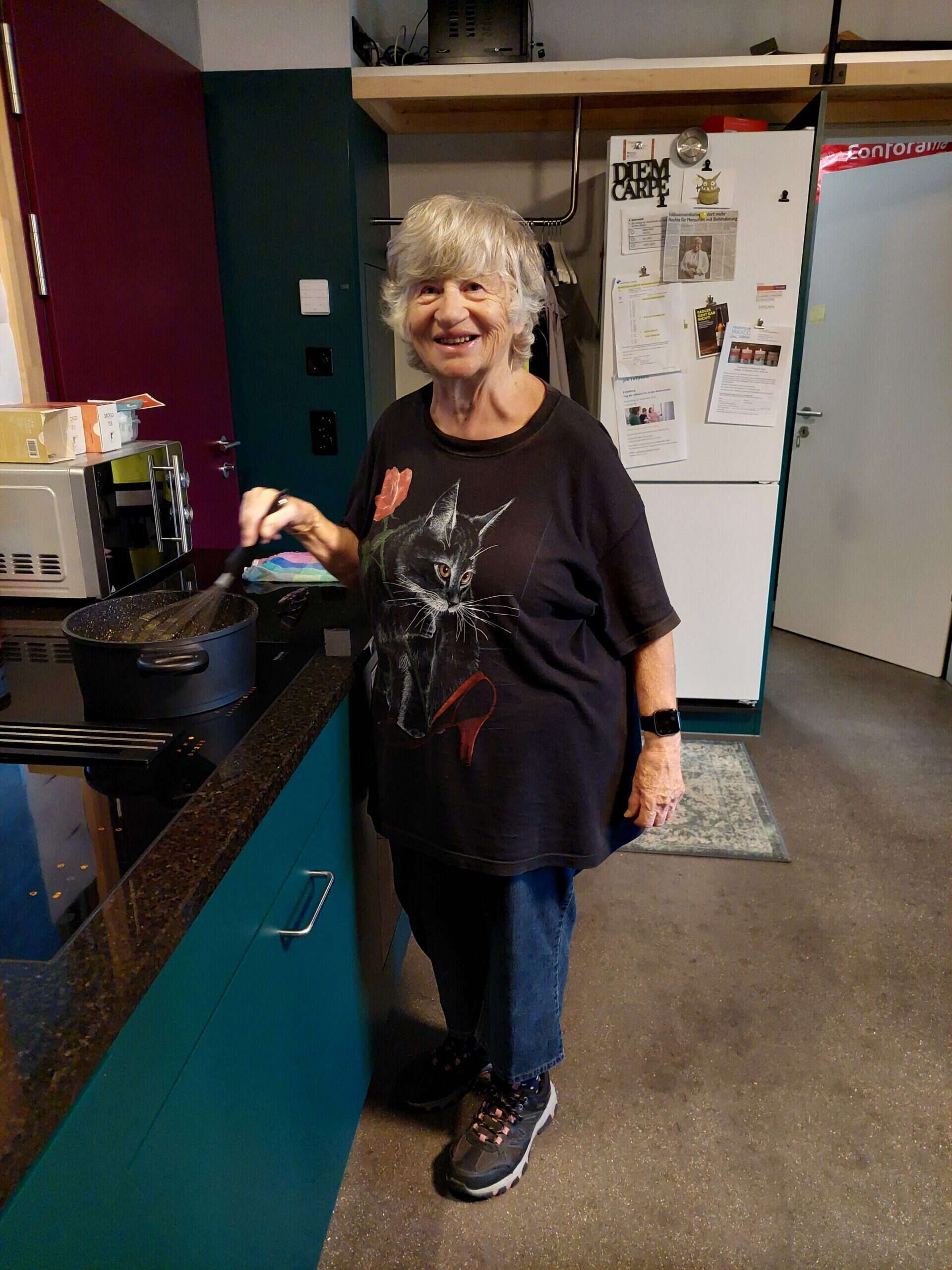 Eine ältere Frau mit grauem Haar steht in einer Küche und lächelt in die Kamera, während sie einen Topf auf dem Herd umrührt - eine warme Szene aus einer tagesstätte für menschen mit behinderung, die tagesstruktur für erwachsene bietet. Sie trägt ein schwarzes T-Shirt mit einer Katzengrafik.