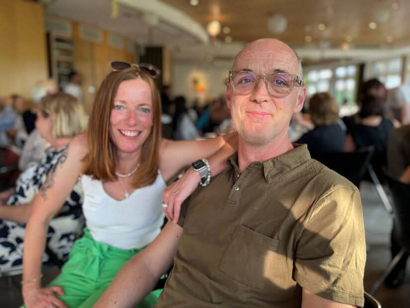 Eine Frau und ein Mann sitzen dicht beieinander und lächeln in die Kamera in einem hell erleuchteten, überfüllten Innenraum.