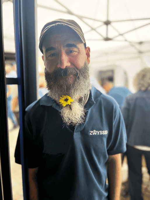 Ein Mann mit grauem Bart und Mütze steht in einem Haus und trägt ein blaues Poloshirt mit der Aufschrift "ZURYSSTIG". Sein Bart ist mit einer gelben Blume gebunden.