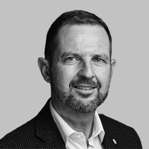 Kopfbild eines Mannes mittleren Alters mit kurzen Haaren und Bart, der eine Anzugsjacke und ein Hemd mit Kragen trägt und in die Kamera lächelt, vor einem schlichten Hintergrund.