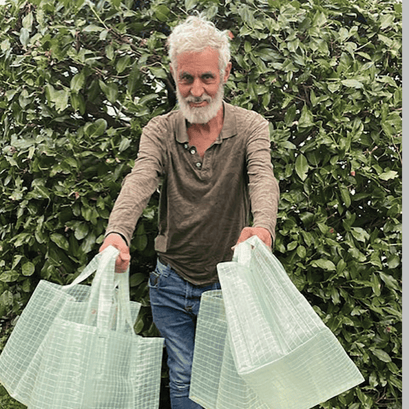 Ein älterer Mann mit grauem Haar und Bart steht vor einem grünen Gebüsch und hält mehrere große grüne Einkaufstüten in beiden Händen, als käme er aus einem Atelier voller kreativer Fundstücke.