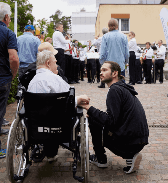 Ein Mann hockt neben einer älteren Person im Rollstuhl und hält ihre Hand, während im Hintergrund eine Gruppe von Musikern im Freien spielt.