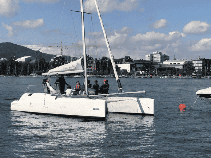 Ein weißer Katamaran mit mehreren Personen an Bord segelt auf einem ruhigen Gewässer, das an die Freiheit und Unterstützung in der Ambulanten Wohnbegleitung erinnert, während im Hintergrund eine Stadtlandschaft und Berge zu sehen sind.