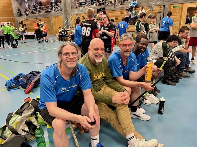 Vier Männer sitzen auf einer Bank in einer Turnhalle, halten Sportgeräte in der Hand, im Hintergrund sind andere Spieler und Zuschauer zu sehen - versammelt, um durch ihre Liebe zum Sport für soziale Projekte zu spenden.