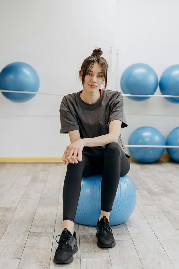 Eine Frau in grauem T-Shirt und schwarzen Leggings sitzt auf einem blauen Gymnastikball in einem Fitnessstudio, in dem spenden soziale projekte den Zugang zu den Geräten unterstützen, mit mehreren anderen blauen Gymnastikbällen im Hintergrund.