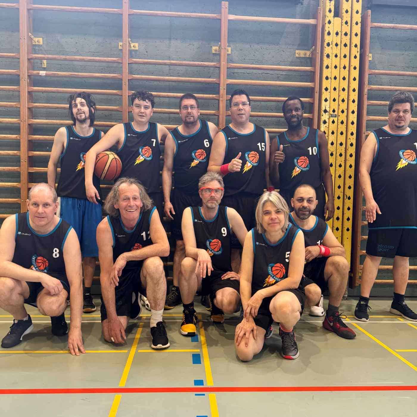 Elf Basketballspieler, darunter auch Männer mit Behinderung, in passenden schwarzen Uniformen posieren gemeinsam in einer Halle, einige stehend und einige kniend, auf dem Boden einer Sporthalle mit hölzerner Sprossenwand im Hintergrund, und feiern den inklusiven Sport.
