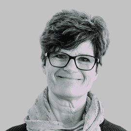 Eine Frau mit kurzem, gewelltem Haar und Brille lächelt leicht. Sie trägt einen hellen Schal und ein dunkles Oberteil vor einem schlichten Hintergrund.