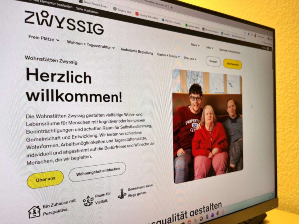Auf einem Computerbildschirm erscheint die Homepage der Wohnstätten Zwyssig mit einem Willkommensgruß und einem Gruppenfoto, auf dem drei Personen auf einer Couch sitzen.