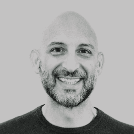 Lächelnder Mann mit Glatze und Bart, der ein dunkles T-Shirt mit Rundhalsausschnitt trägt und vor einem schlichten hellgrauen Hintergrund nach vorne schaut.