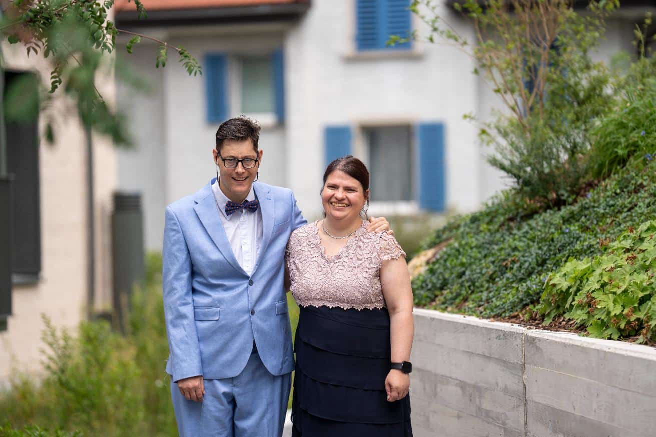 Zwei Personen stehen im Freien nebeneinander und lächeln in die Kamera. Der Mann trägt einen hellblauen Anzug und eine Fliege, die Frau ein kurzärmeliges Kleid mit einem Spitzenoberteil.