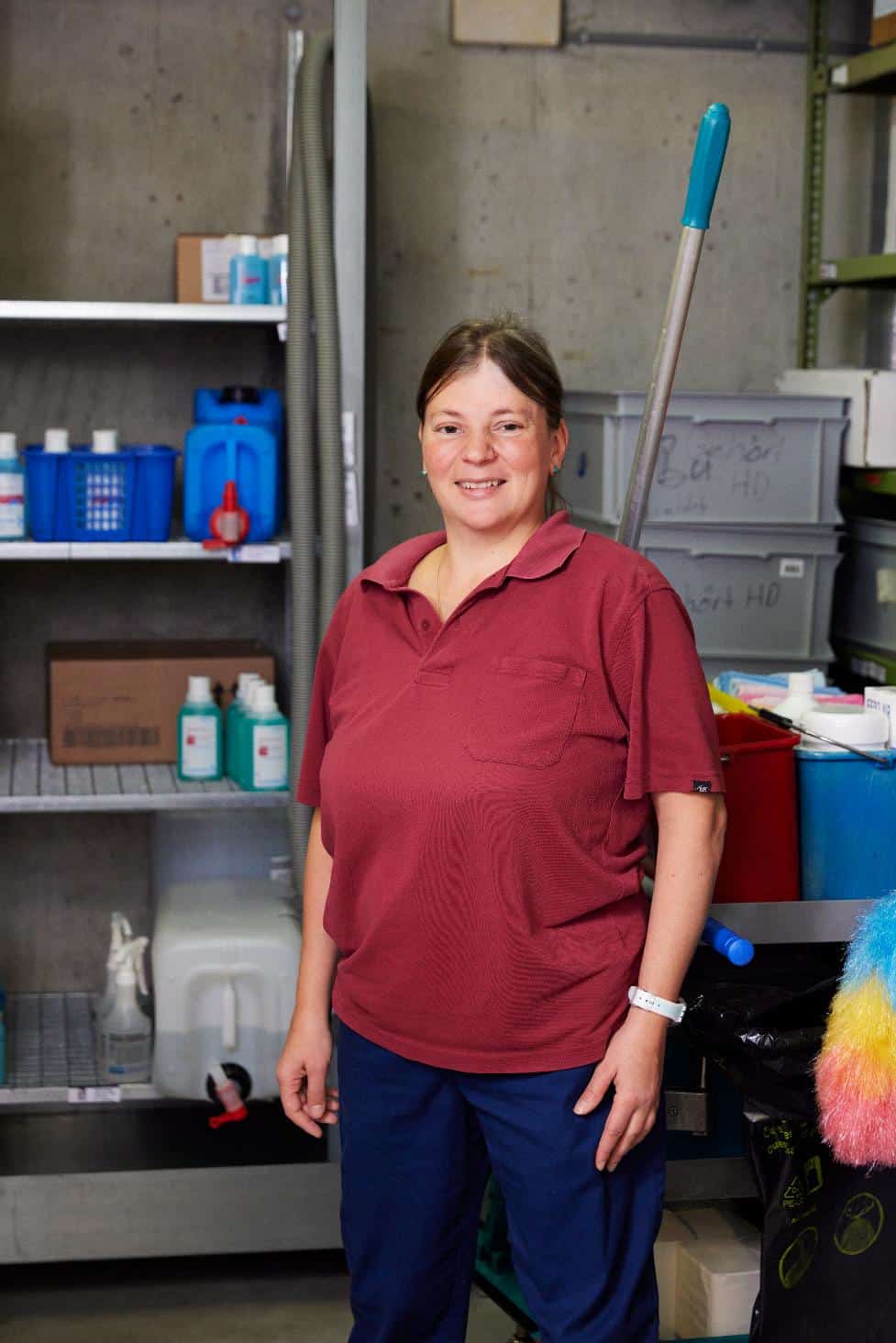 Eine Frau in einem roten Polohemd und einer blauen Hose steht in einem Vorratsraum mit Reinigungsmitteln und -geräten und lächelt in die Kamera.