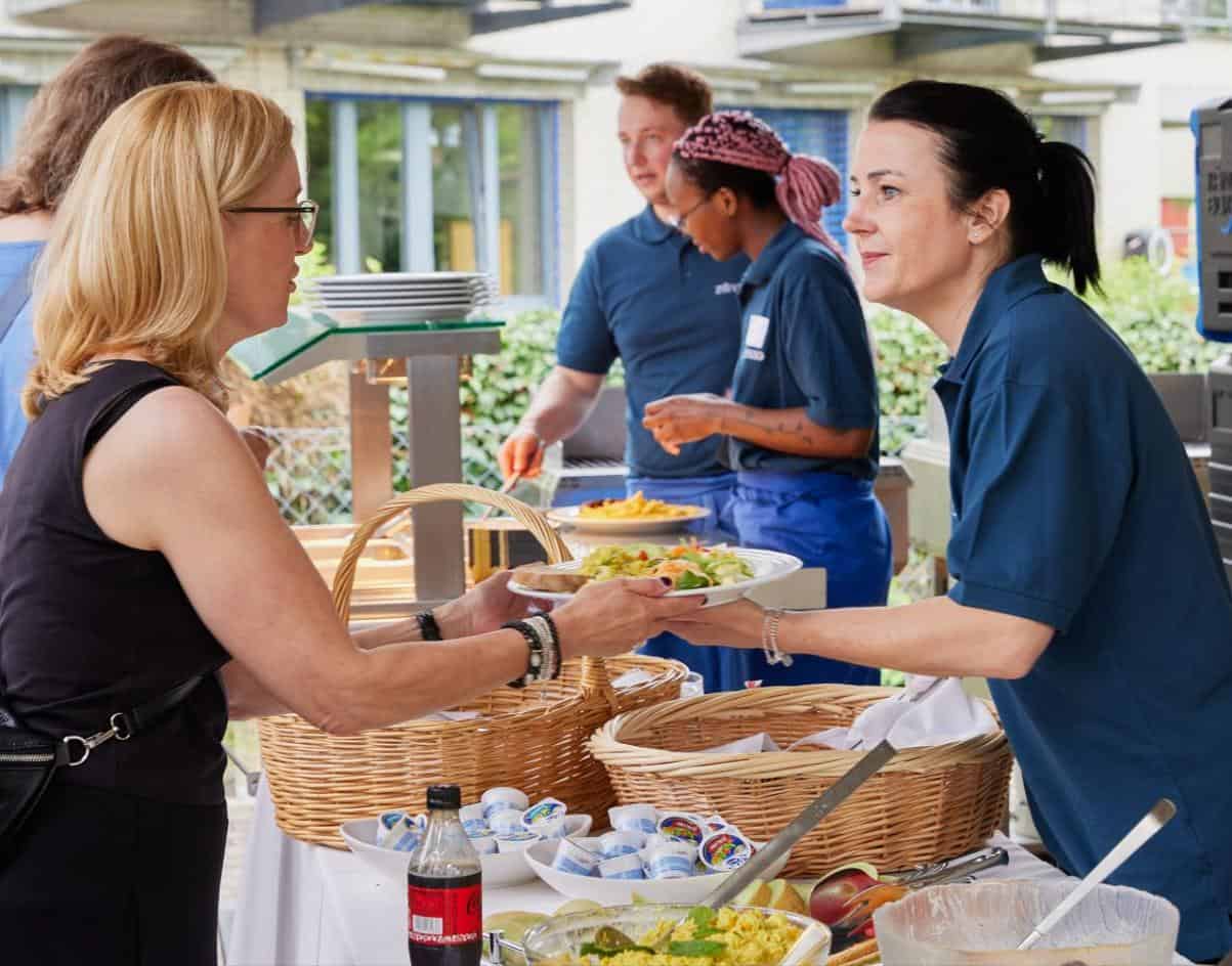 Eine Frau erhält einen Teller mit Essen von einem Kellner an einem Buffet im Freien, auf dem Salate, Brot und Getränke stehen. Im Hintergrund sind weitere Personen zu sehen.