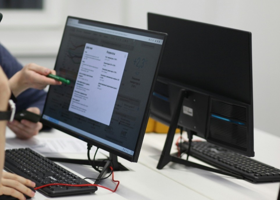 Zwei Personen arbeiten an nebeneinander stehenden Desktop-Computern auf einem weißen Schreibtisch, wobei die eine auf einen Monitor mit einem Dropdown-Menü zeigt.