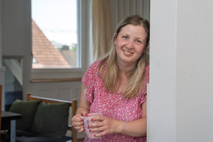 Eine Frau in einem rosa geblümten Hemd steht in einem Haus, lehnt an einer Wand, hält eine Tasse in der Hand und lächelt in die Kamera.